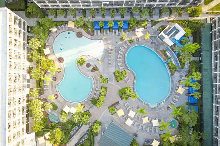 Aerial view of two resort pools surrounded by lounge chairs, tropical plants, and buildings
