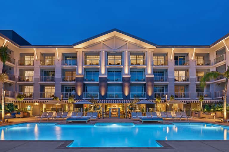 Modern resort exterior at dusk with illuminated pool, multi-story building, and loungers reflected in calm water