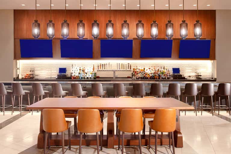 Modern hotel bar with orange chairs, wooden accent wall, and blue-lit shelving with bottles