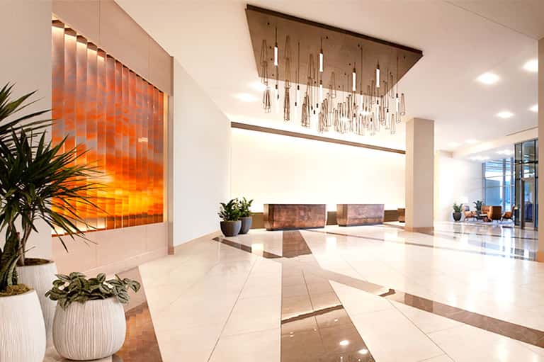 Modern hotel lobby with polished marble floors, orange accent wall, suspended lighting fixture, and potted plants