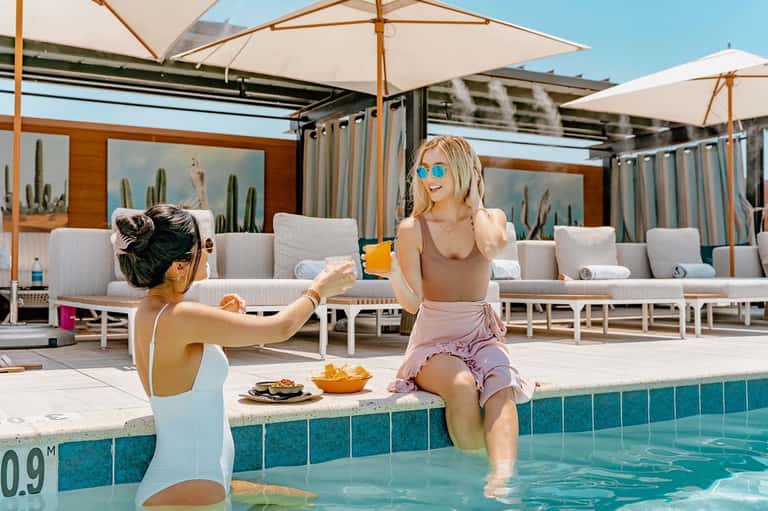 Two women enjoying drinks by resort pool under white umbrellas with modern facilities