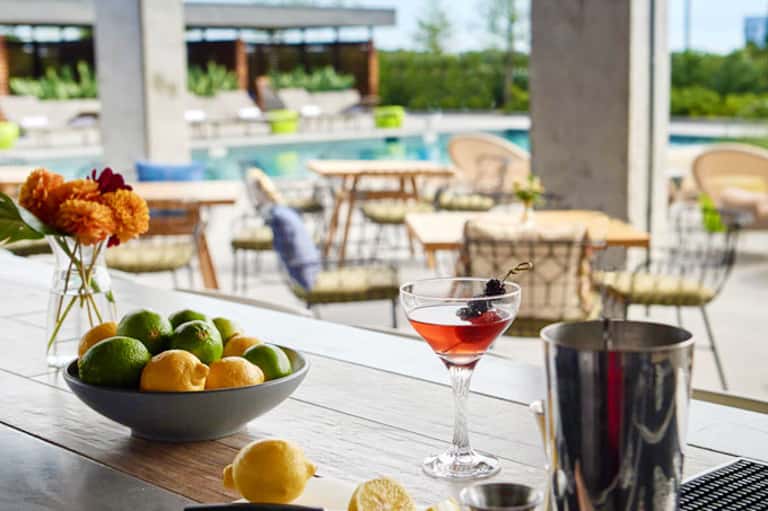 Outdoor poolside bar setup with citrus fruits, cocktail, and lounge chairs overlooking modern resort grounds