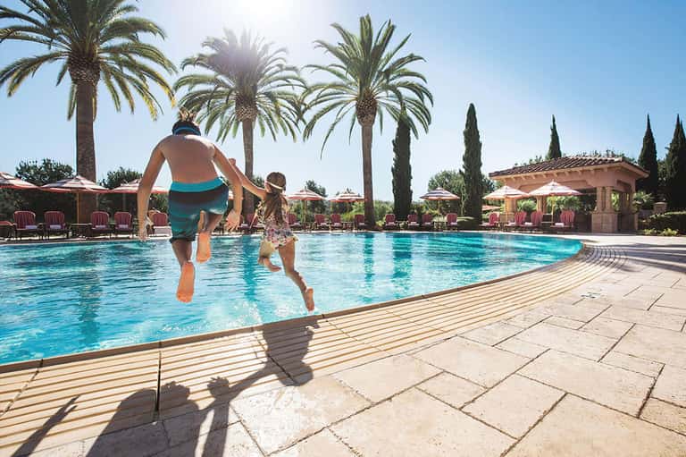 Father and child running into resort pool with palm trees and lounge chairs under Mediterranean sun