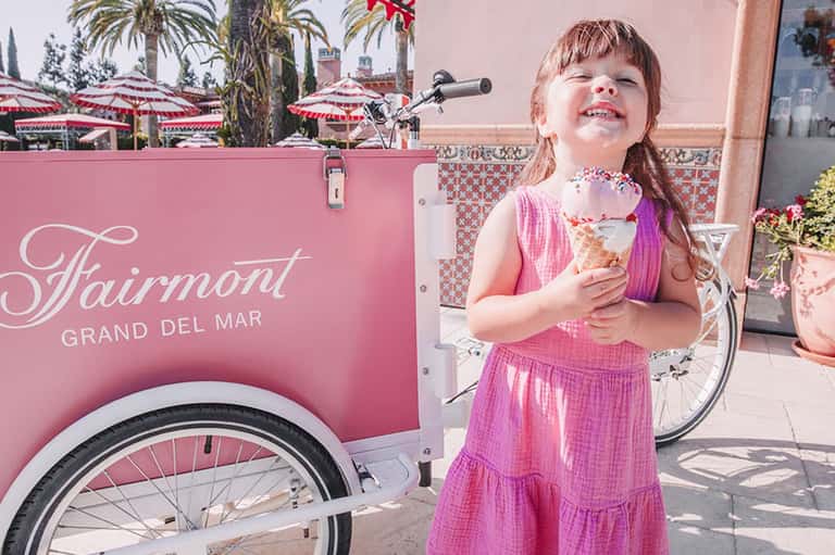 Young girl in pink dress holding ice cream cone next to Fairmont Grand Del Mar pink ice cream cart