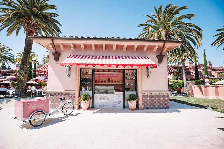 Pink kiosk with red and white striped awning and matching bicycle in palm tree-lined plaza