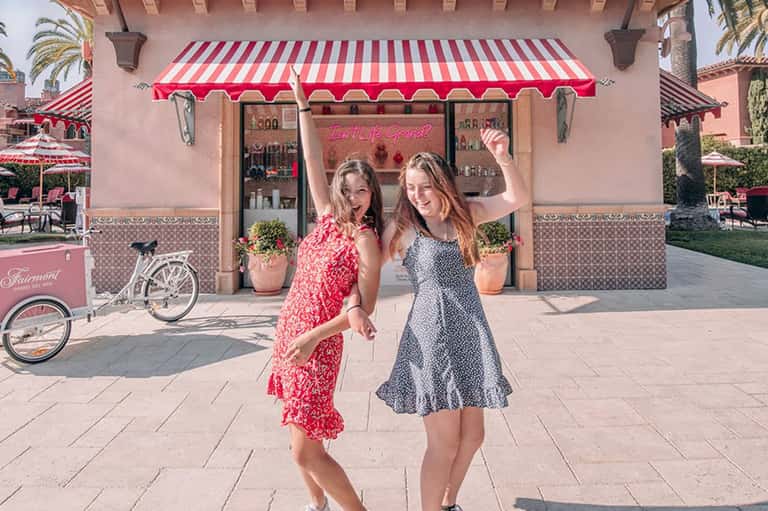 Two women posing outside a pink storefront with red and white striped awning in a resort courtyard
