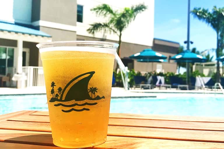 Yellow beverage with shark logo design in cup by resort pool with palm trees and blue umbrellas