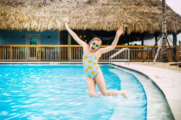 Child in yellow swimsuit jumping in pool with thatched-roof beach house structure
