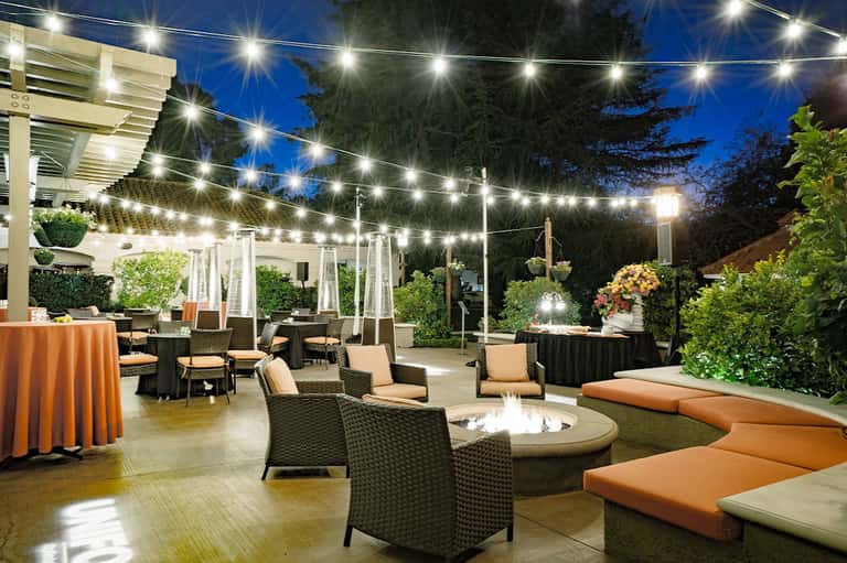 Elegant outdoor event space with string lights, fire pit, orange linens, and modern seating at dusk