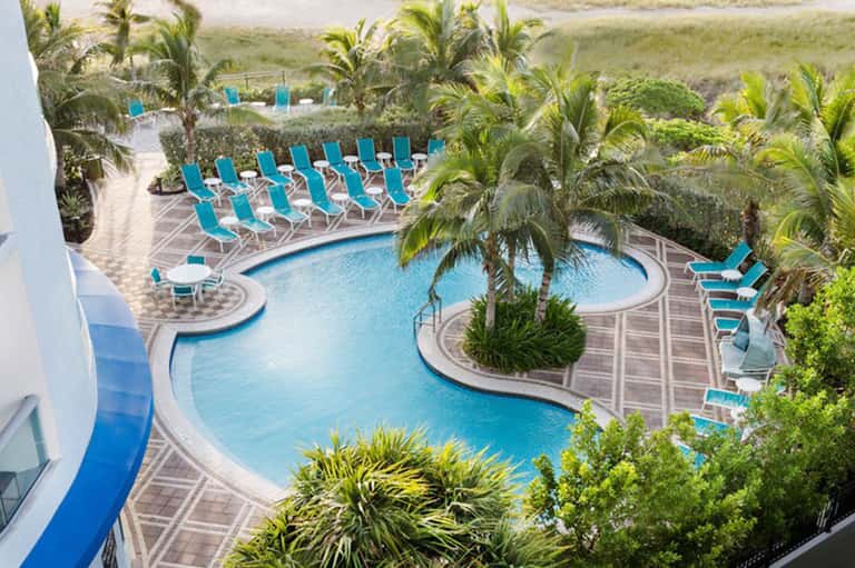 Aerial view of curved resort pool with blue loungers, palm trees, and beach access