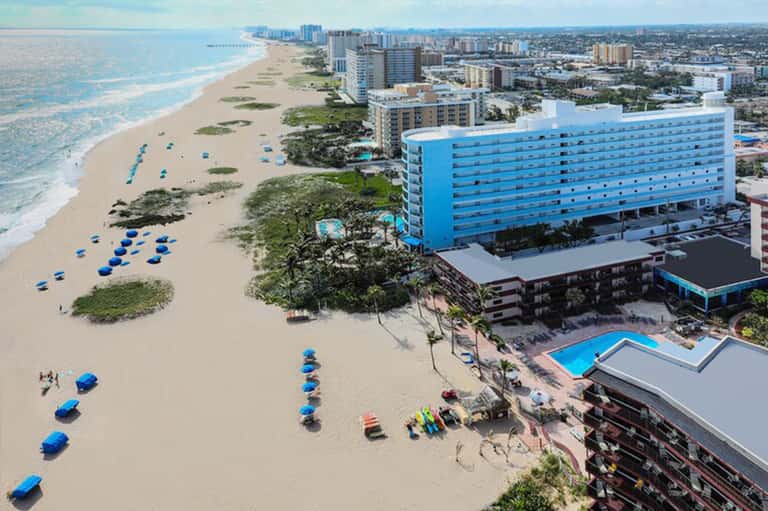 Aerial view of beachfront resort with bright blue building, sandy beach with blue umbrellas, and coastal cityscape