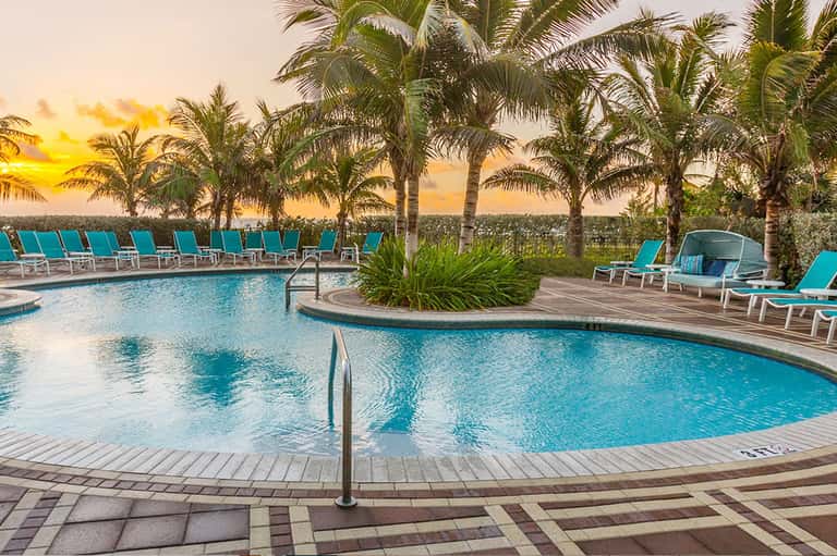 Resort pool at sunset with turquoise water, palm trees, lounge chairs, and colorful sky