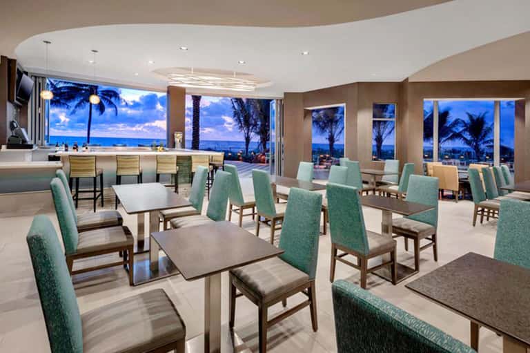 Upscale beachfront dining room with turquoise chairs, tables, and panoramic ocean views at dusk