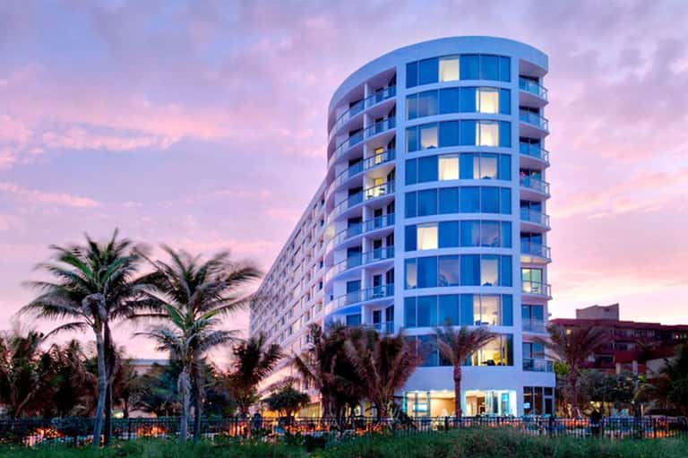 Modern blue curved building at dusk with palm trees and illuminated windows