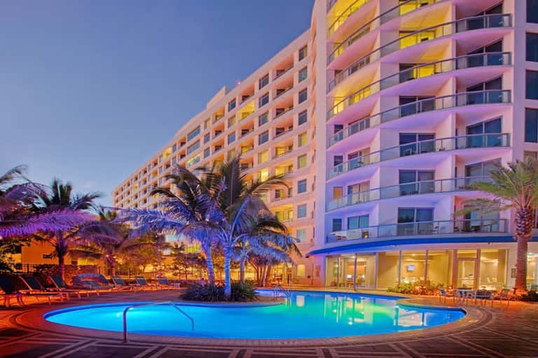 Pink and orange resort building with illuminated pool at dusk, featuring palm trees and lounge chairs