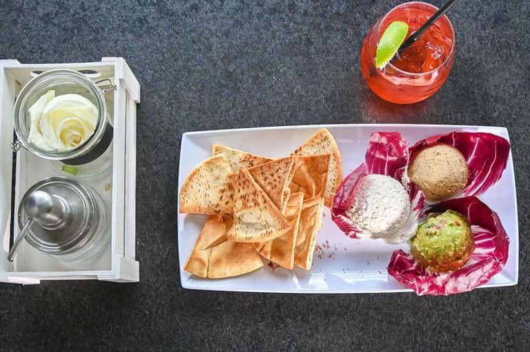 Overhead view of cocktails, appetizers with hummus and guacamole, and crispy pita chips on a dark table