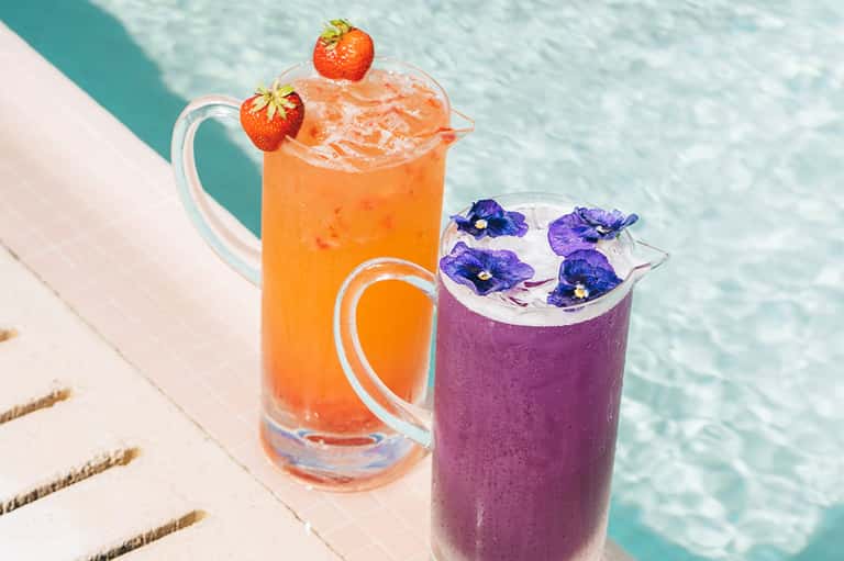 Two colorful cocktails by a turquoise pool: orange strawberry pitcher and purple flower-garnished drink