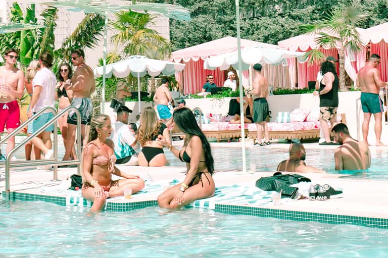 Busy resort pool with guests swimming, pink and white cabanas, palm trees, and lounge chairs