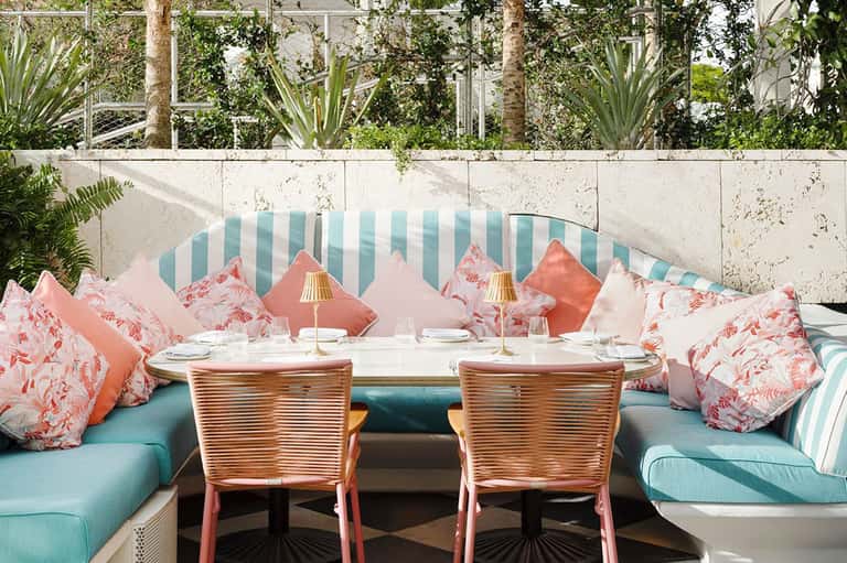 Outdoor dining area with turquoise striped banquette seating, coral and pink patterned pillows, and tropical garden backdrop