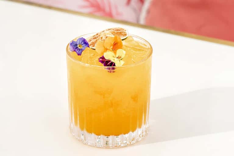 Golden cocktail with edible flowers and ice in crystal glass on white surface
