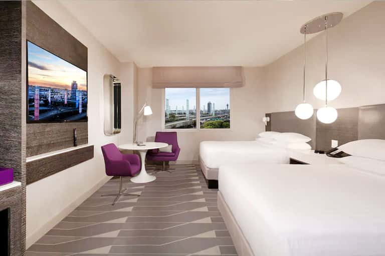 Modern hotel bedroom with two beds, city views, purple chairs, and pendant lighting