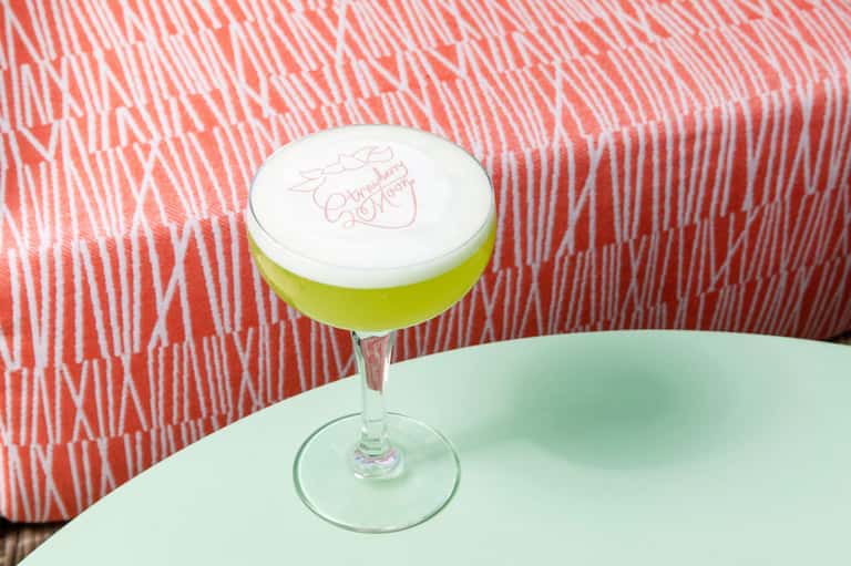 Bright green cocktail with white foam top and pink text on stem glass against red patterned background
