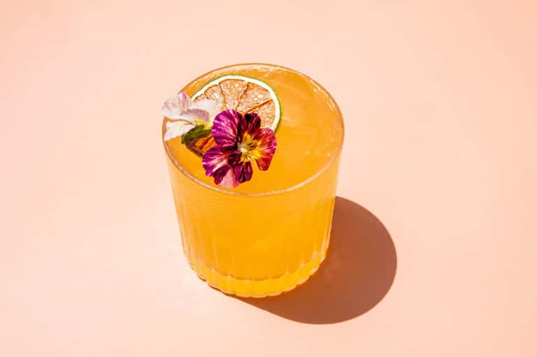 Yellow cocktail with orange slice and purple flower garnish on pale pink background