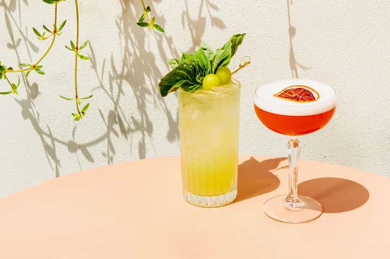 Two colorful cocktails on a peach table with mint garnish and citrus slice against white wall
