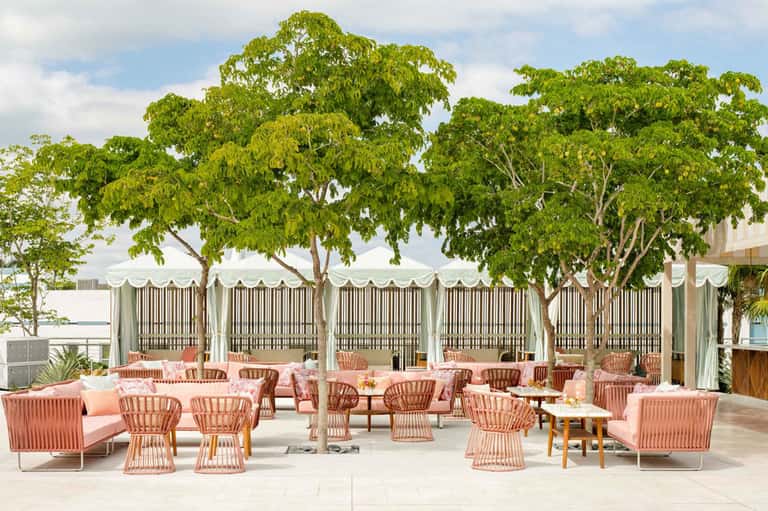 Upscale outdoor terrace with coral lounge seating, shade structures, and mature trees overlooking water
