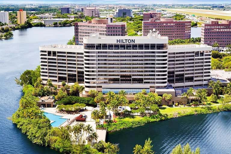 Aerial view of Hilton hotel on waterfront with pool, palm trees, and surrounding cityscape