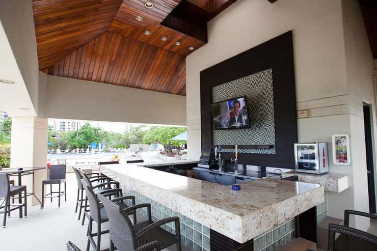 Modern outdoor bar with granite countertop, black tile accent wall, TV, and covered patio seating
