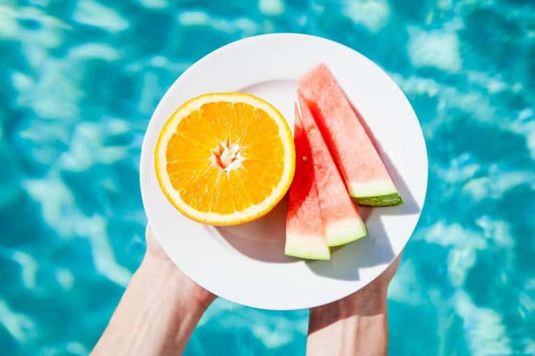Hands holding plate of fresh fruit including orange slice and watermelon above sparkling pool water