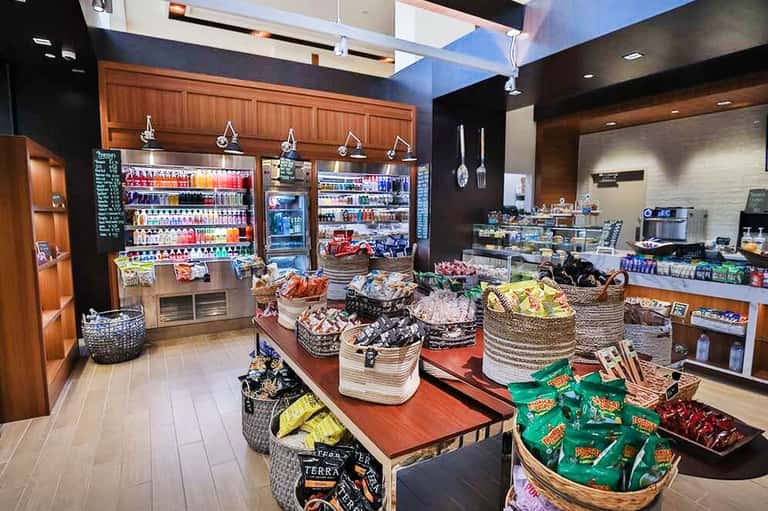 Modern resort convenience store interior with refrigerated beverage cases, snack displays in woven baskets, and checkout counter