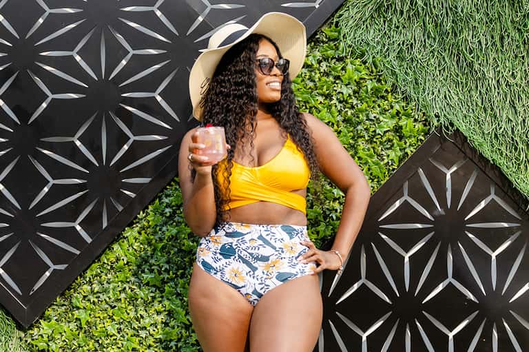 Woman in yellow bikini top and patterned swim shorts posing against decorative wall