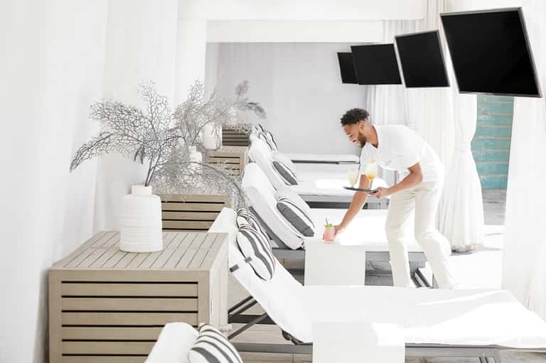 Modern spa lounge with white loungers, potted plants, and staff member serving beverages