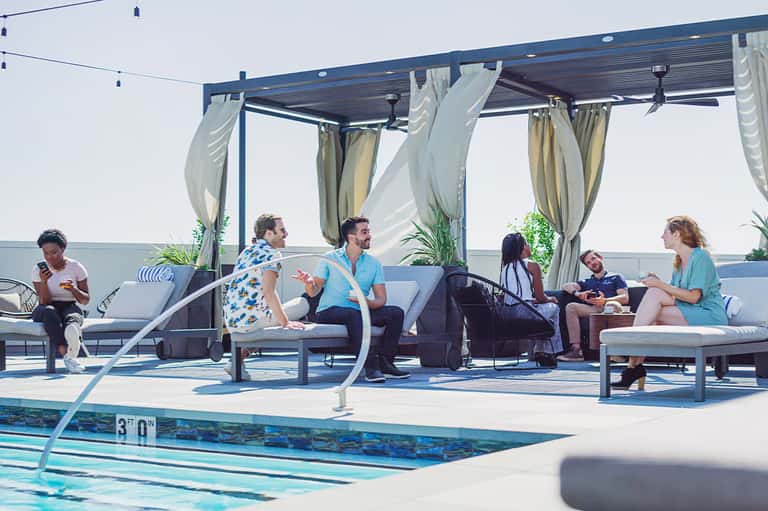 Waterside lounge with pergola, draped curtains, modern furniture, and people relaxing by lap pool