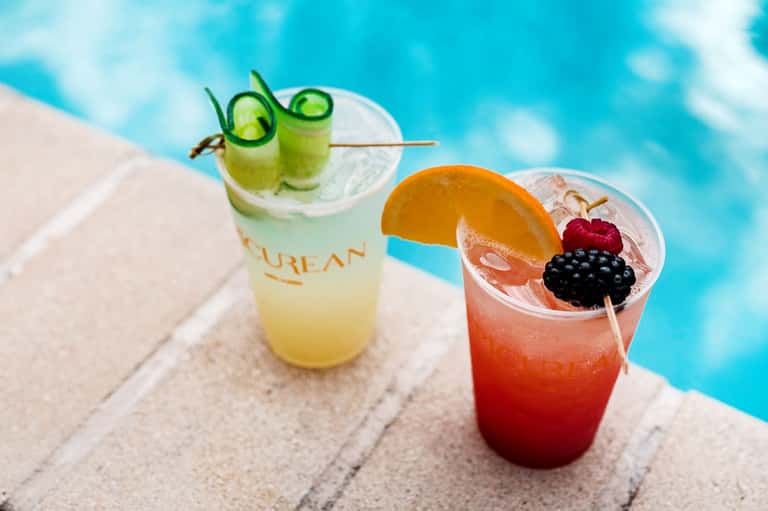 Two colorful cocktails by pool edge with cucumber and berry garnishes