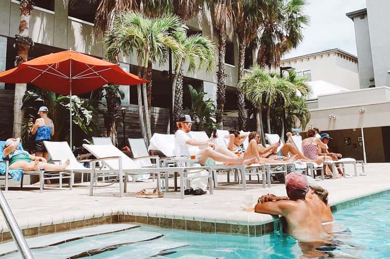 Resort courtyard with guests lounging on white chairs, red umbrella, and palm trees surrounding pool