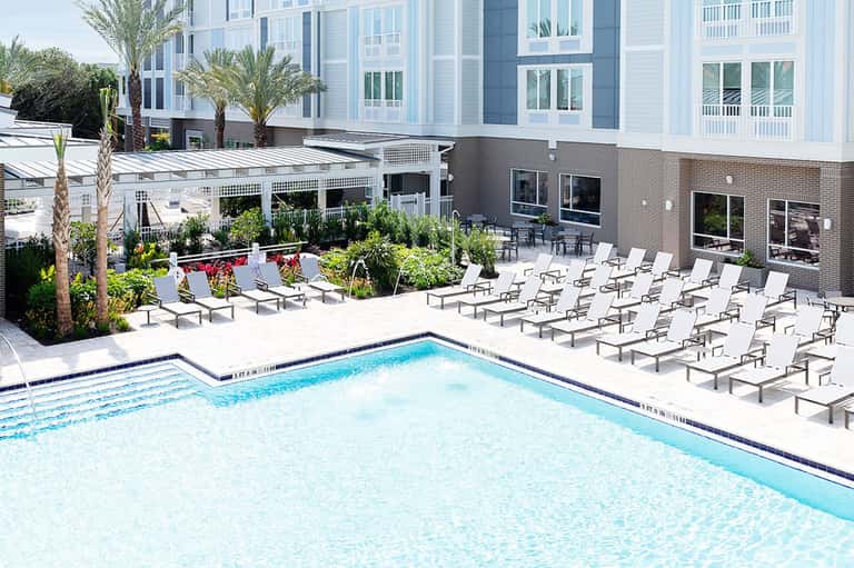 Resort swimming pool with lounge chairs, palm trees, and modern brick building