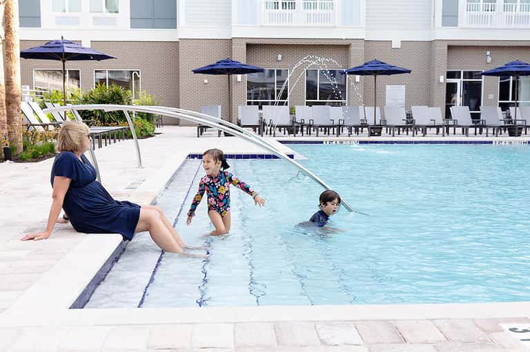 Woman and children swimming in outdoor pool with lounge chairs and blue umbrellas under bright sun
