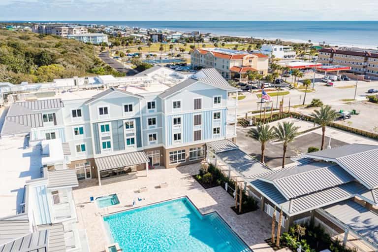 Aerial view of beachfront resort with pool, white buildings, palm trees, and ocean beyond