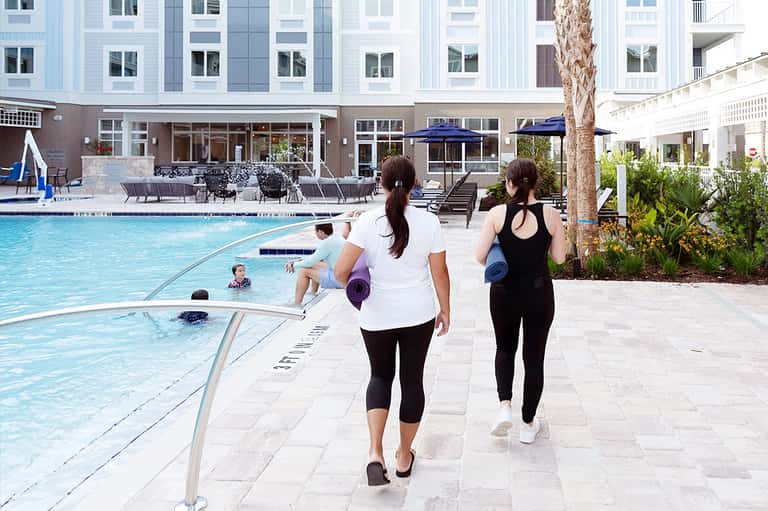 Two women jogging along resort pool deck with turquoise water, lounge chairs, and multi-story building with palm trees