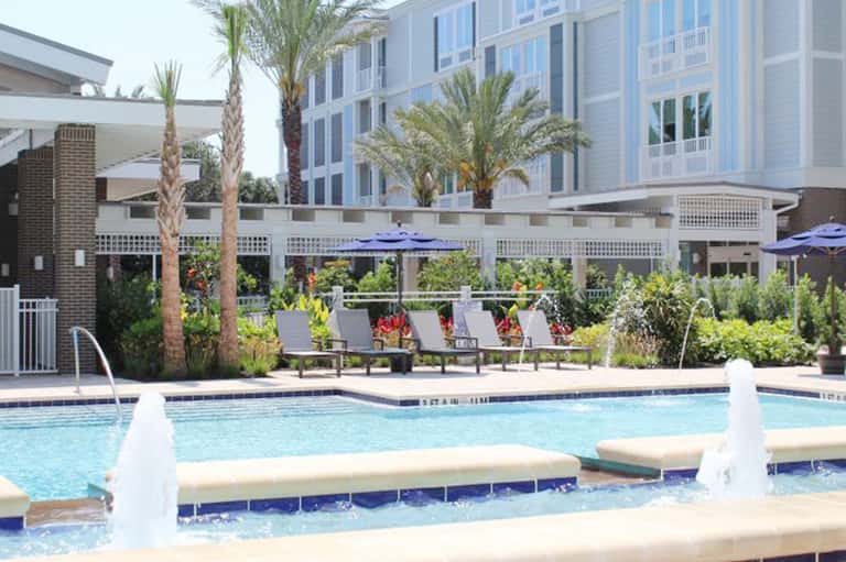 Resort pool area with lounge chairs, blue umbrellas, palm trees, and contemporary building backdrop