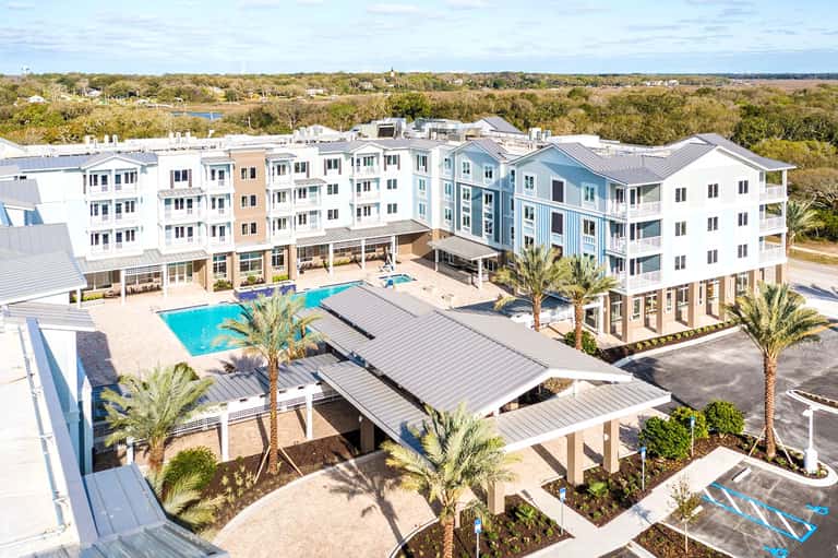 Aerial view of modern coastal resort with blue pool, palm trees, and multi-story white buildings surrounded by green landscape