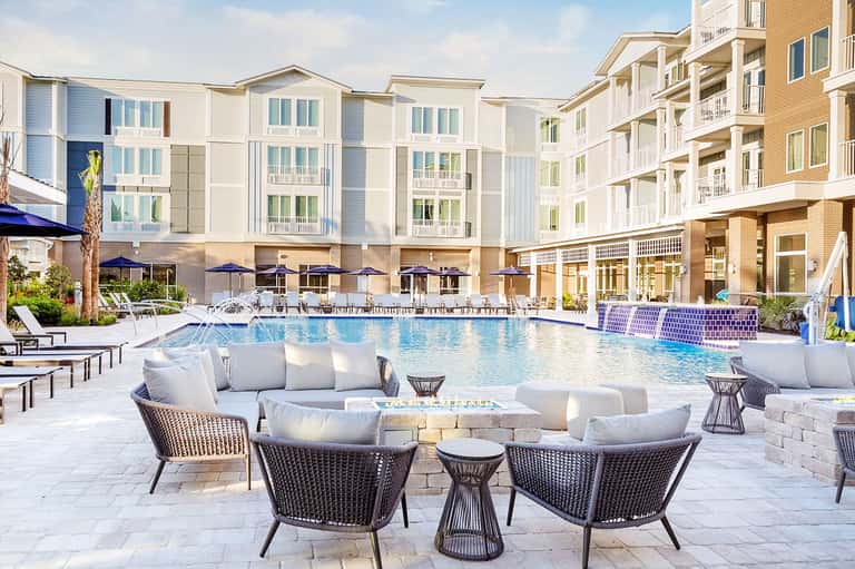 Luxury resort pool area with modern white buildings, lounge seating, and blue umbrellas