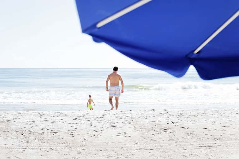 Adult and child wading in ocean waves under a large blue beach umbrella