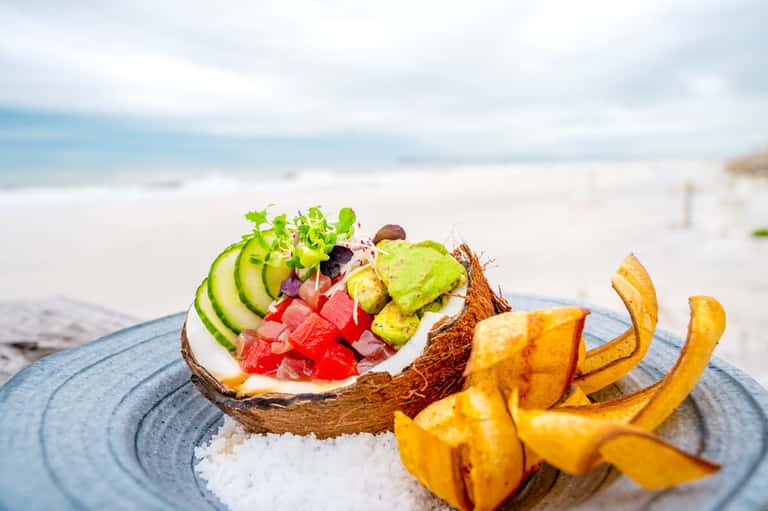 Beachside salad bowl with fresh vegetables and tropical fruit served on white sand near ocean