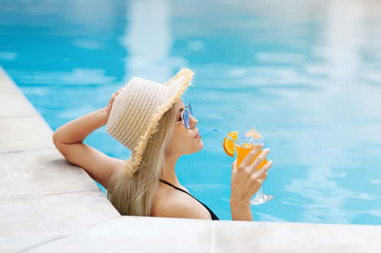 Woman relaxing in resort pool wearing hat and sunglasses, holding orange cocktail