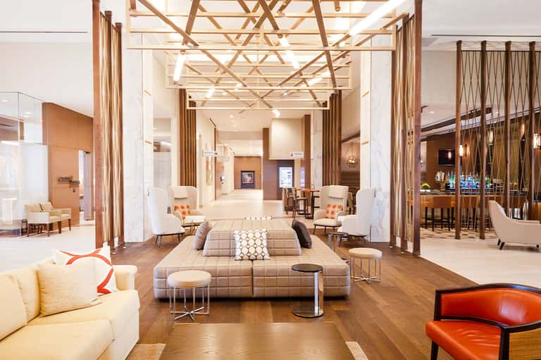 Modern hotel lobby with neutral tones, geometric ceiling, lounge seating, and bar area