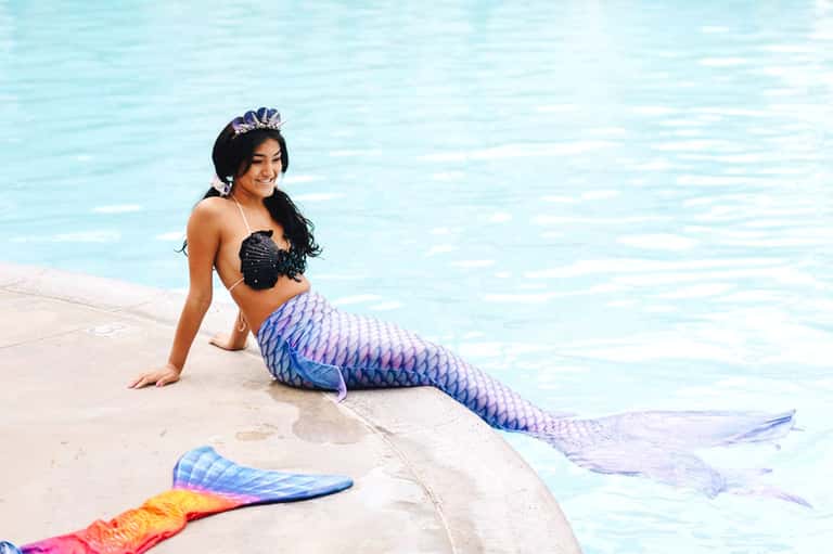 Woman in mermaid tail costume posing by turquoise pool with colorful tail fins nearby